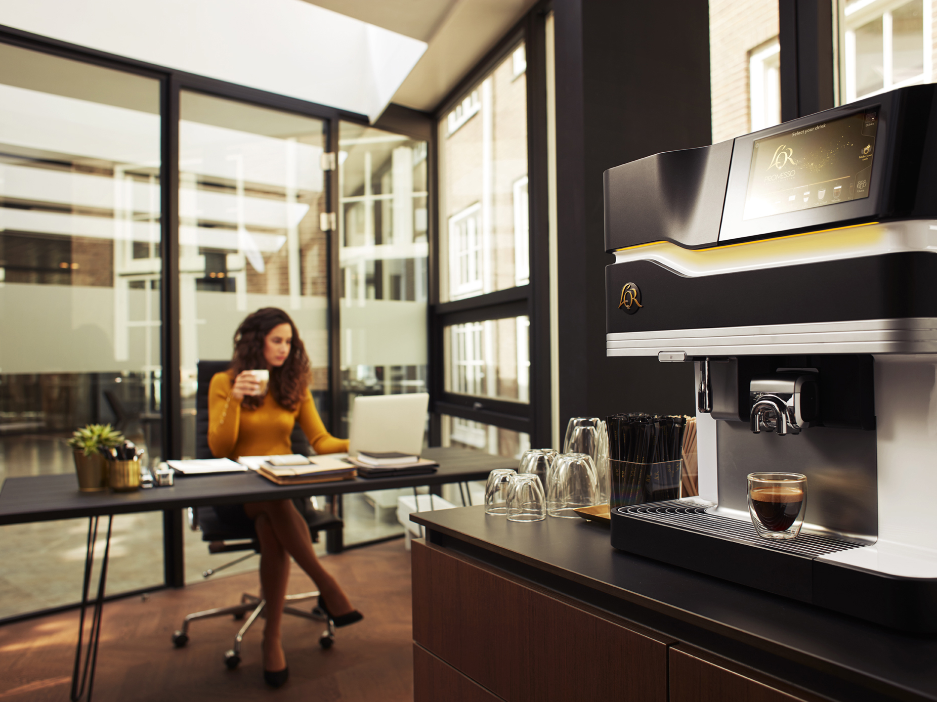 A woman drinking a glass cup of L'OR coffee on a table with a laptop, with a coffee machine
